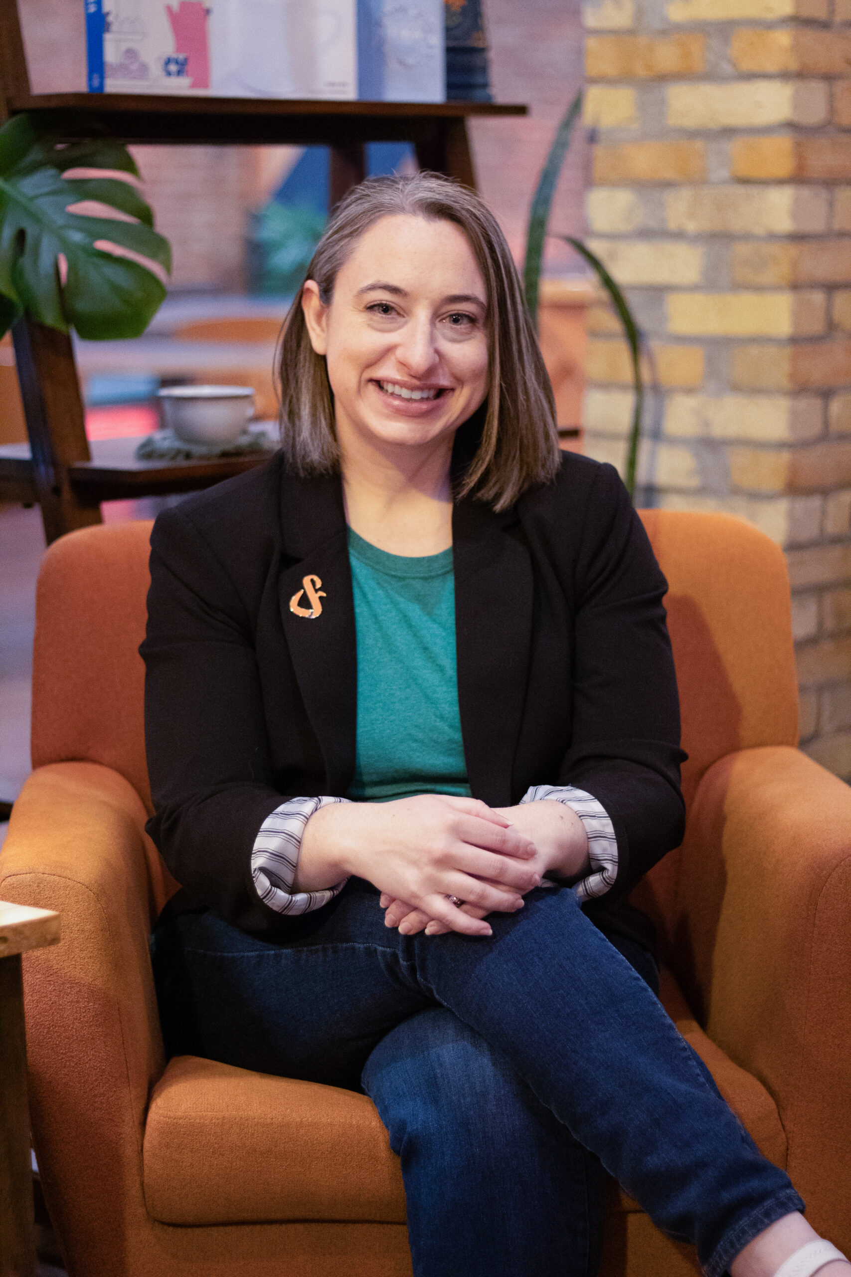 Carrie Jorgensen sits in an orange chair, wearing a jacket with a Lost&Found pin.