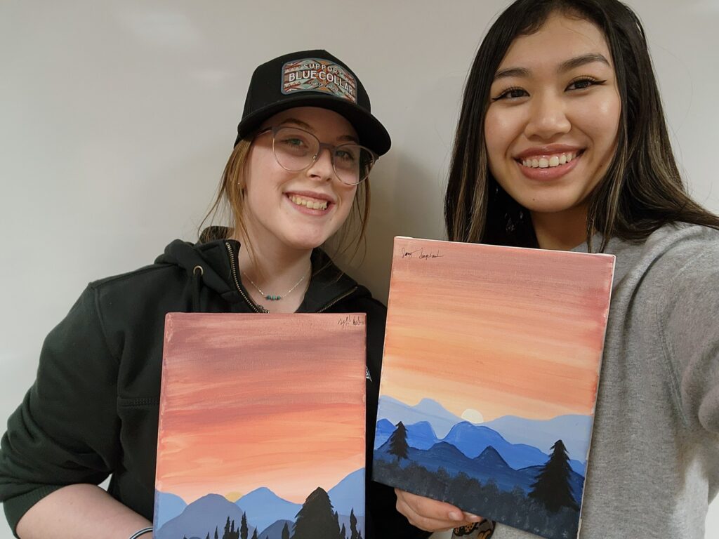 Jenny Sengchanh, right, and her mentee, Kaylah R., share art they've created. The art is paintings of mountains, trees, and a pink/orange sky.